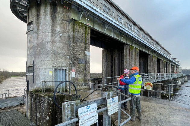 Parteen Weir - large concrete structure - standing within and above the River Shannon. Doherty is wearing a hi-vis jacket with ESB written on it. Lacchia is wearing a hat and red coat while looking towards the river. Signs in the foreground state - ESB Fisheries Conservation. 