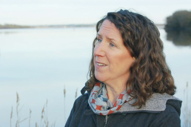 McGoff standing on the banks of a large body of water - looking towards an interviewer - wearing a warm coat and scarf. 