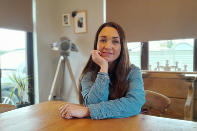 Kayleigh Payne, a 36 year old white woman with long brown hair sits at a kitchen table. She has a slight smile.  One hand is on the table and her face rests on the other hand as she looks at the camera.