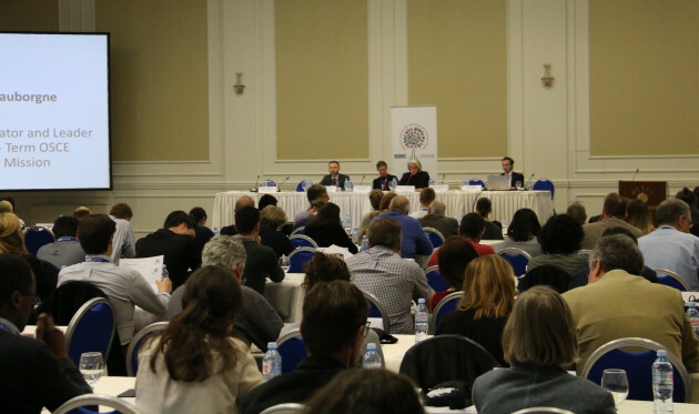 A large group of election observers sitting in rows listening to a panel of four experts of the OSCE. 