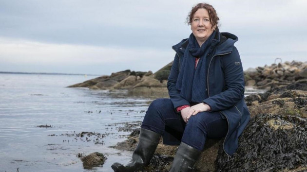 Dr Róisín Nash smiling in a navy jacket sitting on a large rock beside the sea