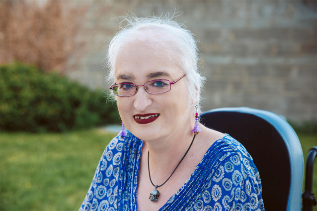 Selina Bonnie - wearing a blue patterned top, necklace and earrings - sitting in a garden. The top of her wheelchair is visible.