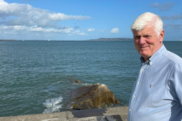 Jones stands in front of Dublin Bay with a view of Howth in the background. He is wearing a blue shirt.
