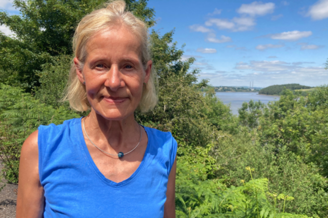 Dubsky wearing a blue top and necklace standing in front of trees with the sea in the background.