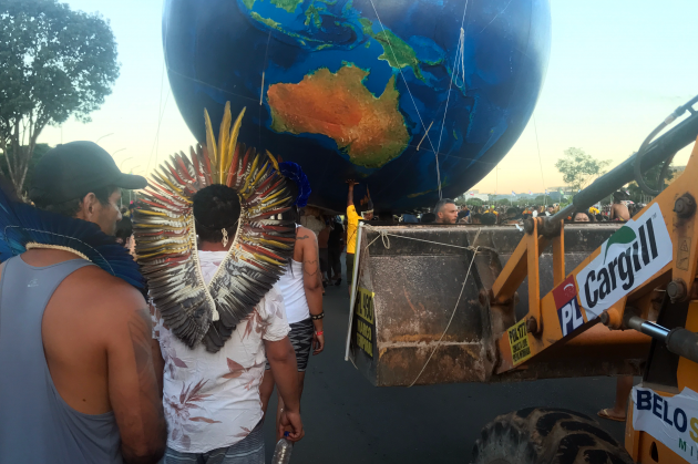 A number of people with native headdresses walk beside a digger with Cargill signs attached to it. A large globe is being held by protesters in the background.