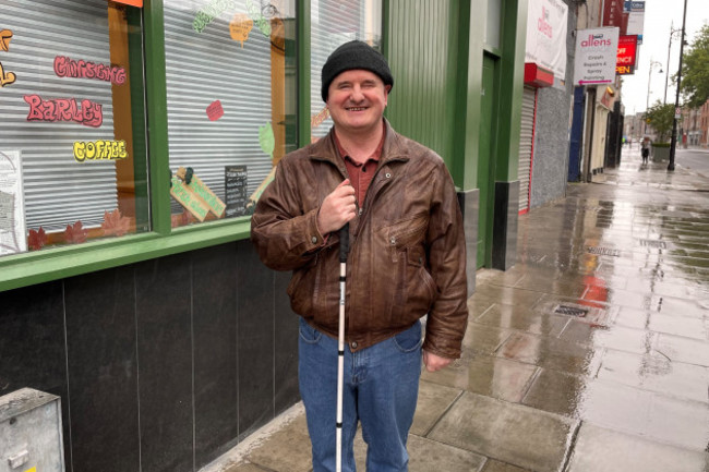 Robbie Sinnott - wearing a cap, brown leather jacket and jeans, carrying a cane - standing on a wet street outside a coffee shop painted in green. 