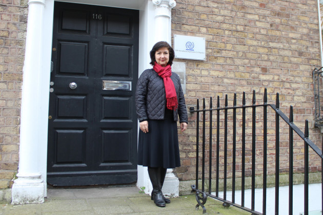 Zuzana Vatralova, wearing all black with a read scarf, stands at the entrance to the IOM offices in Dublin. The sign beside the door reads 