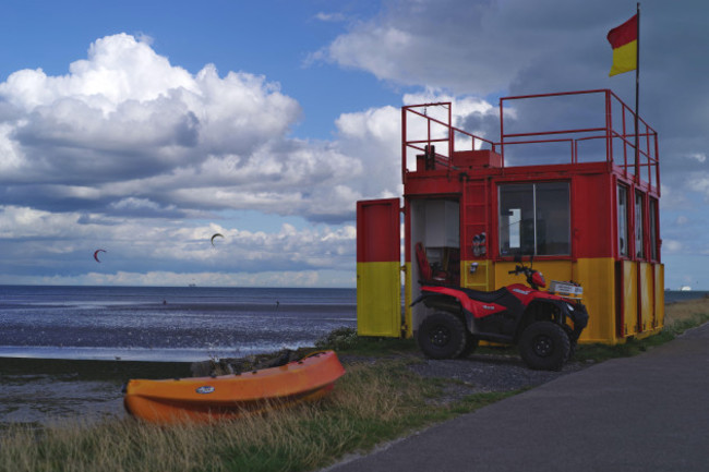 lifeguards-on-duty-in-red-and-yellow-hut-on-the-dollymount-strand-beach-in-dublin-ireland-paragliders-and-the-cloudy-summer-sky-in-the-background