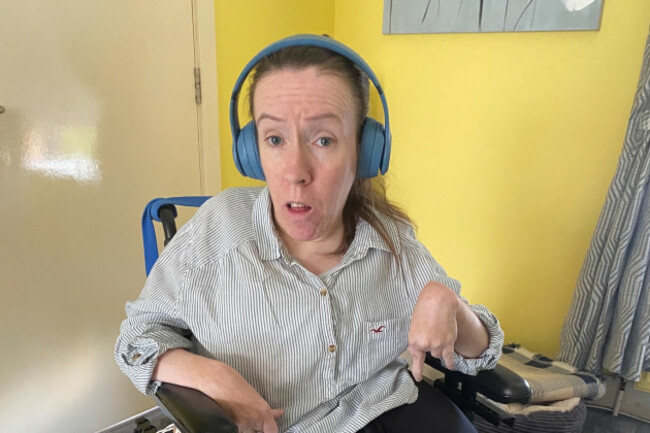 Shelly, a wheelchair user, wearing a pale striped shirt sitting and headphones while holding the movement controls of her wheelchair with her left hand.