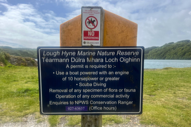 Signs at the edge of Lough Hyne. The top says - No horse riding - and the bottom says - Lough Hyne Marine Reserve. Téarmann Dúlra Mhara Loch Oighinn. A permit is required to - Use a boat powered with an engine of 10 horsepower or greater, Scuba Diving, Removal of any specimen of flora or fauna, Operation of any commercial activity. Enquiries to NPWS Conservation Ranger.