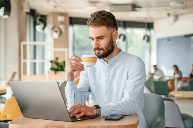 with-cup-of-drink-young-man-is-sitting-in-the-cafe-and-working-by-using-laptop