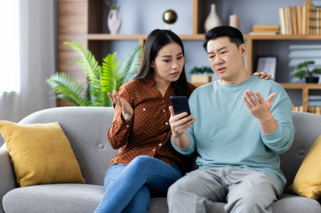 asian-couple-sitting-on-couch-looking-at-phone-with-confused-expressions-discussing-something-important-image-shows-emotional-interaction-communication-relationship-dynamics-technology-use