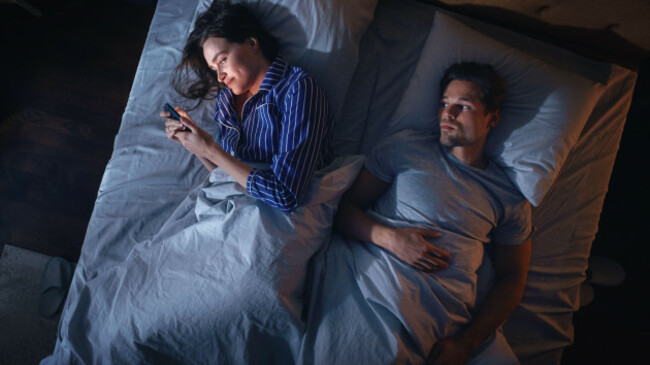top-view-young-woman-uses-smartphone-in-bed-at-night-when-her-male-partner-trying-to-fall-asleep-beside-couple-fight-argue-addictive-world-of-social-media-doom-scrolling-fake-news