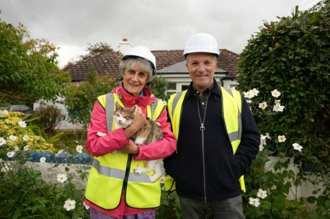 Heat My Home homeowner Sylvia Thompson and presenter Kieran McCarthy with PPE in front of old house Landscape with cat