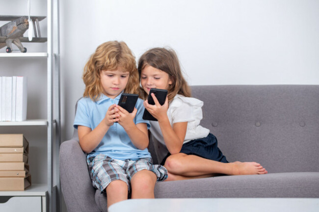 two-kids-using-online-app-on-mobile-phone-making-call-watching-content-browsing-internet-playing-video-games-sitting-on-couch-in-at-home-alone