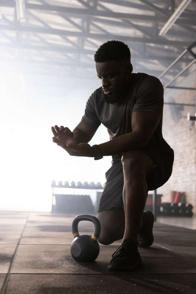 african-american-man-kneeling-chalking-hands-in-warehouse-gym-with-kettlebell-in-dark-shirt-lifter-athlete-focus-dust-industrial-silhouette-bac