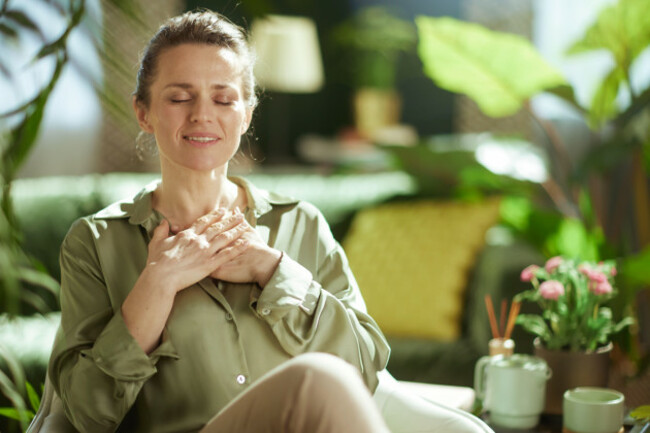 a-woman-finds-tranquility-and-self-compassion-resting-her-hands-on-her-chest-with-a-gentle-smile-in-her-cozy-plant-filled-living-room