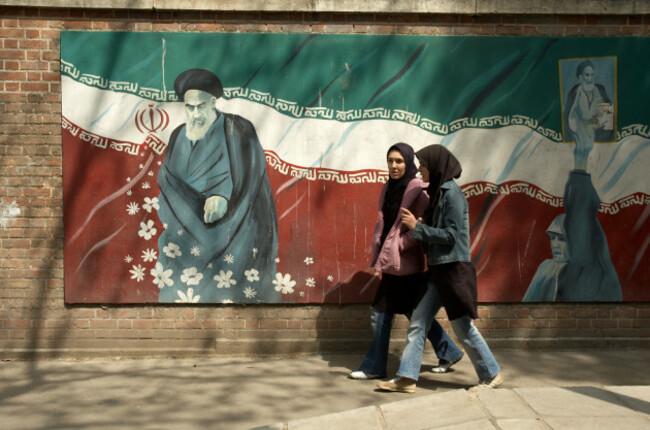 two-woman-wearing-islamic-uniforms-walking-in-front-of-the-painting-of-ayatollah-khomeini-and-the-iranian-flag-on-outside-walls