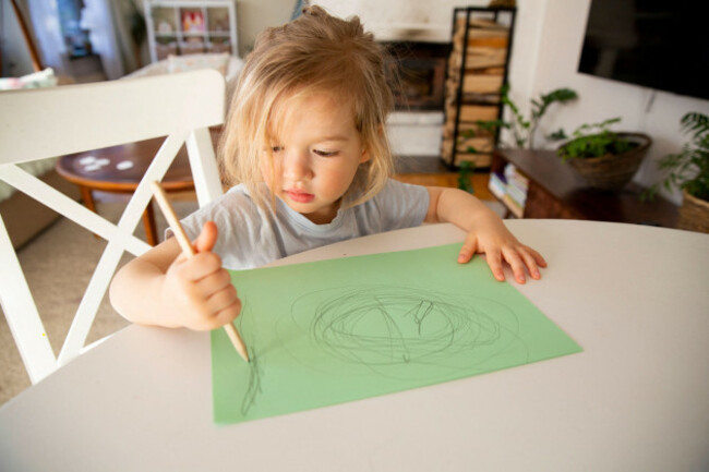 child-with-long-hair-drawing-scribbles-on-green-paper-with-wooden-pencil-sitting-at-table-in-cozy-home-creative-early-development-and-analog-play