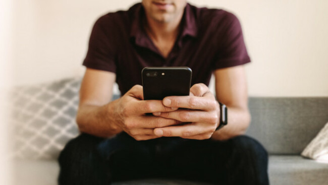 close-up-of-man-working-on-mobile-phone-sitting-on-sofa-man-operating-mobile-phone-with-two-hands