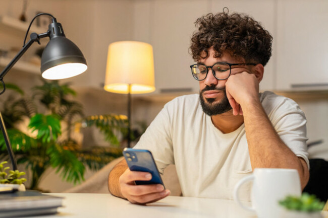 arabian-man-at-home-scrolls-smartphone-news-feels-bored-stressed-negative-expression-sighs-wants-rest-egyptian-guy-at-table-doomscrolls-late-evenin