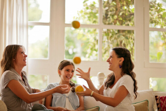 two-sisters-and-their-mother-laughing-together-on-sofa-while-the-older-sister-juggles-fruit