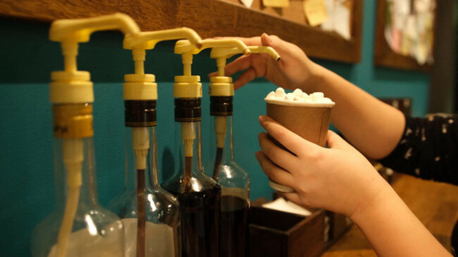 author-coffee-bartender-before-serving-pours-caramel-syrup-topping-into-finished-drink-in-paper-cup-background-blurred