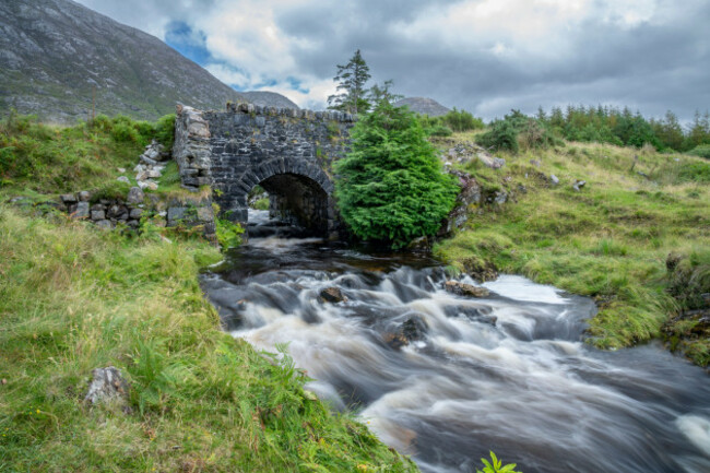 this-image-captures-a-charming-stone-bridge-arching-over-a-cascading-mountain-stream-set-against-the-backdrop-of-the-maumturk-mountain-range-in-conne