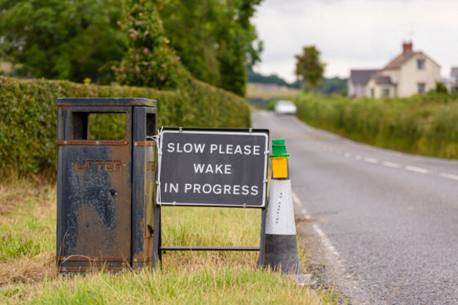 sign-with-a-black-background-and-the-words-slow-please-wake-in-progress-with-a-black-and-white-traffic-cone-and-warning-light-by-the-side-of-a-roa